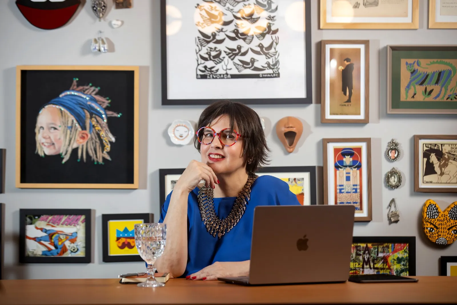 Dani Ramos à mesa de trabalho, diante de uma parede de quadros de arte popular